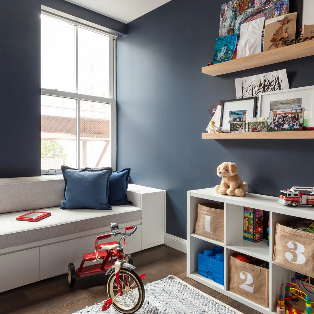 A blue-walled play corner contains white cube storage with numbered fabric bins, a gray window bench, wood picture ledges and a red tricycle.