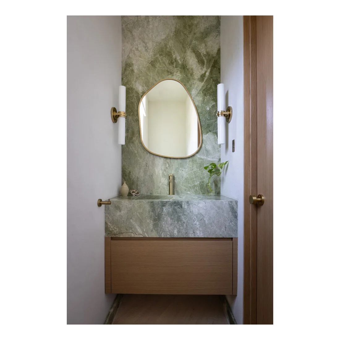 A narrow powder room has a green veined basin counter and tall matching wall panel above a wood drawer, with an irregular mirror and slim white wall lights.