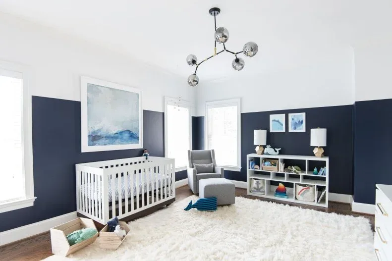 Blue-and-white nursery with white crib, gray adult chair, open storage, pale rug and blue framed artwork.
