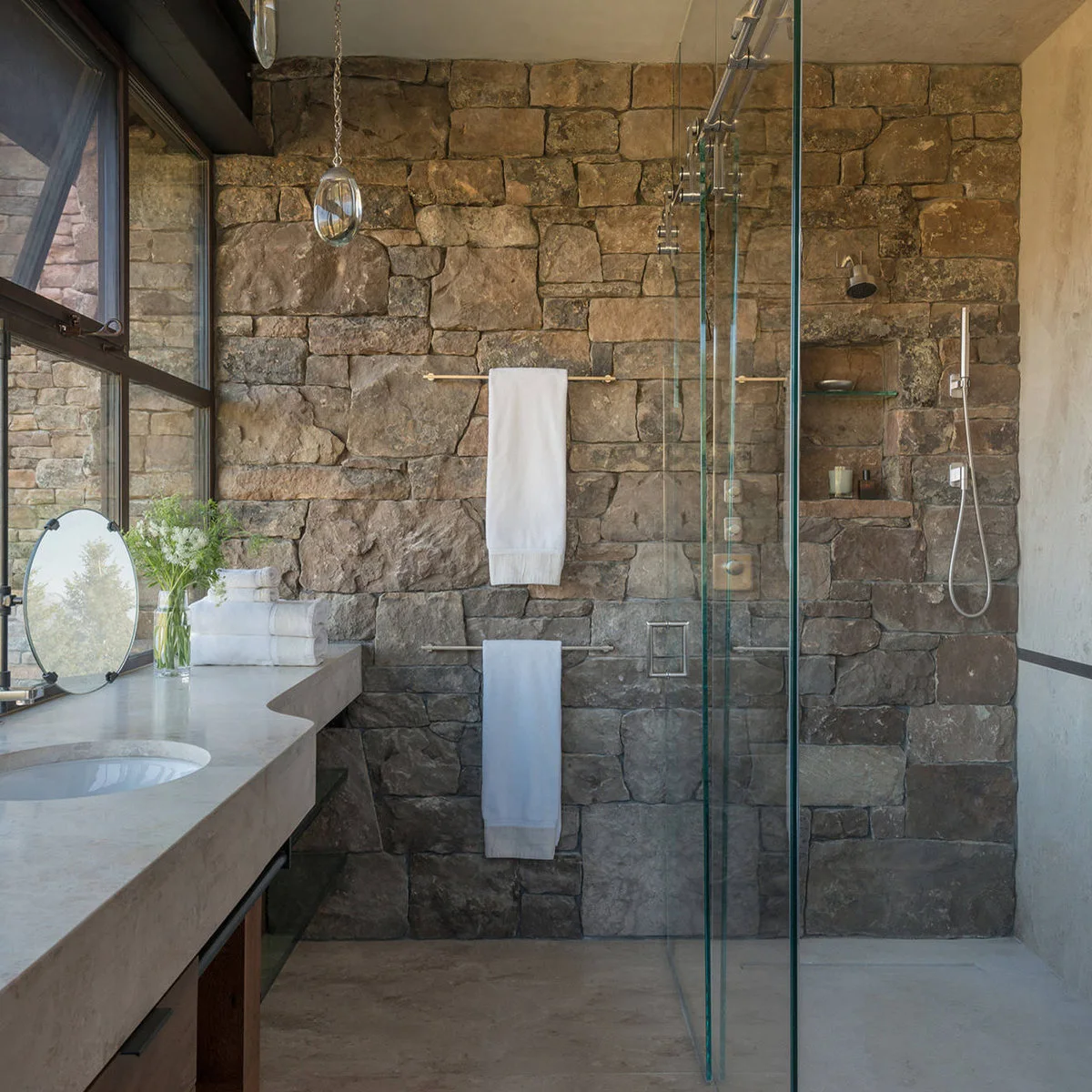 A glass shower against a rough block wall that continues behind towel rails, beside a long pale countertop and black-framed windows.