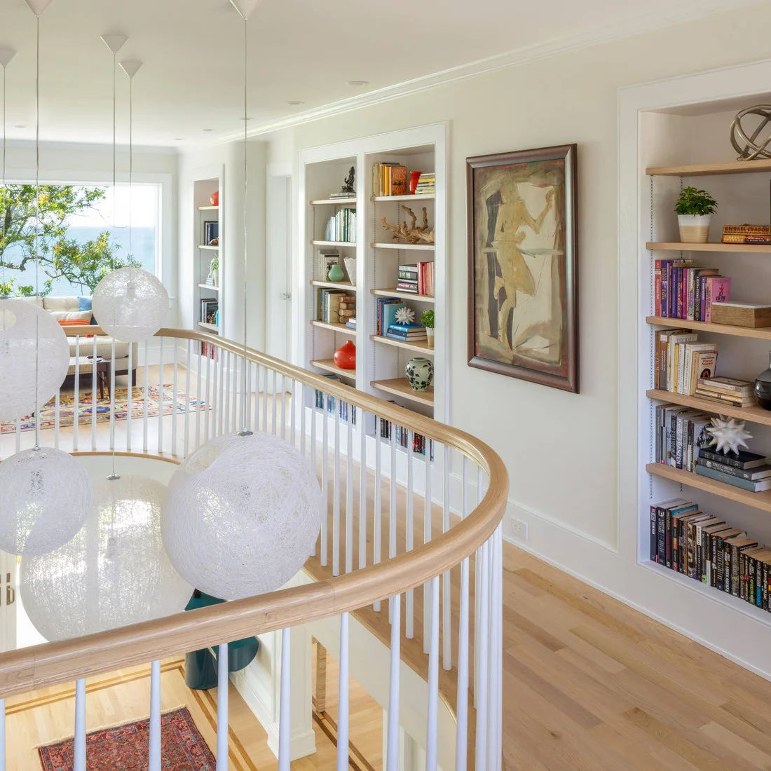 Recessed book shelves with pale wood ledges line a bright upper landing beside a curved white stair rail and large globe pendants.