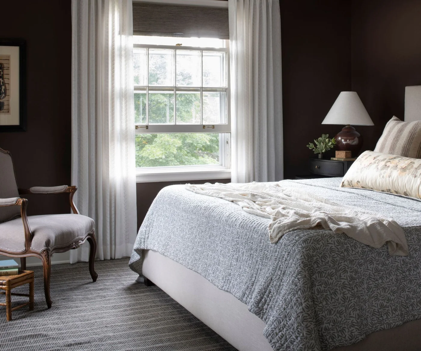 Chocolate brown bedroom walls beside white curtains and pale patterned bedding with a wooden armchair.