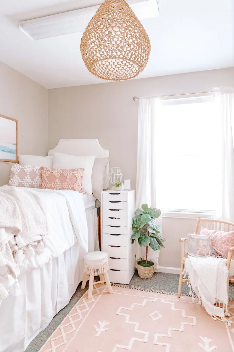 Boho student bedroom with white bedding, pink patterned pillows and rug, white bedside drawers, woven hanging shade and wooden chair beside the window.