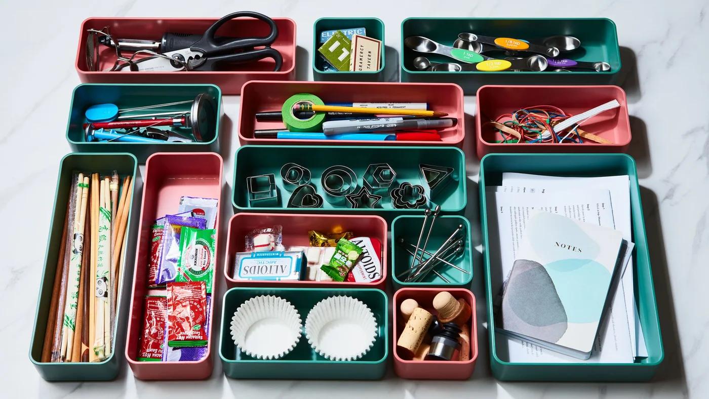 Coral and green modular trays containing stationery, scissors, rubber bands, notes and small kitchen accessories