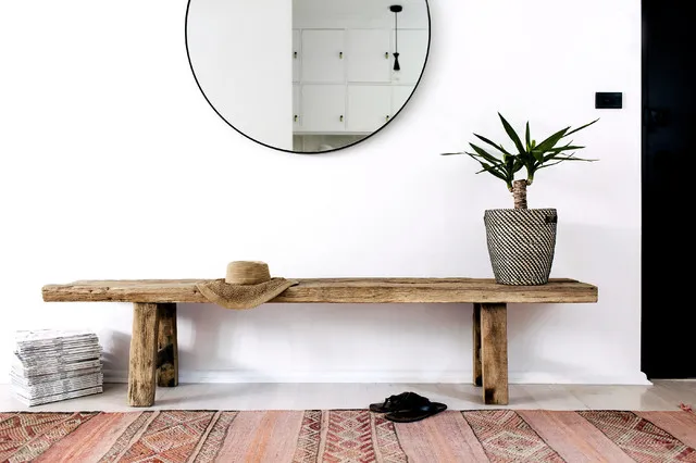 A rough wooden bench sits beneath a round black mirror against a white wall beside a leafy plant and pink patterned rug