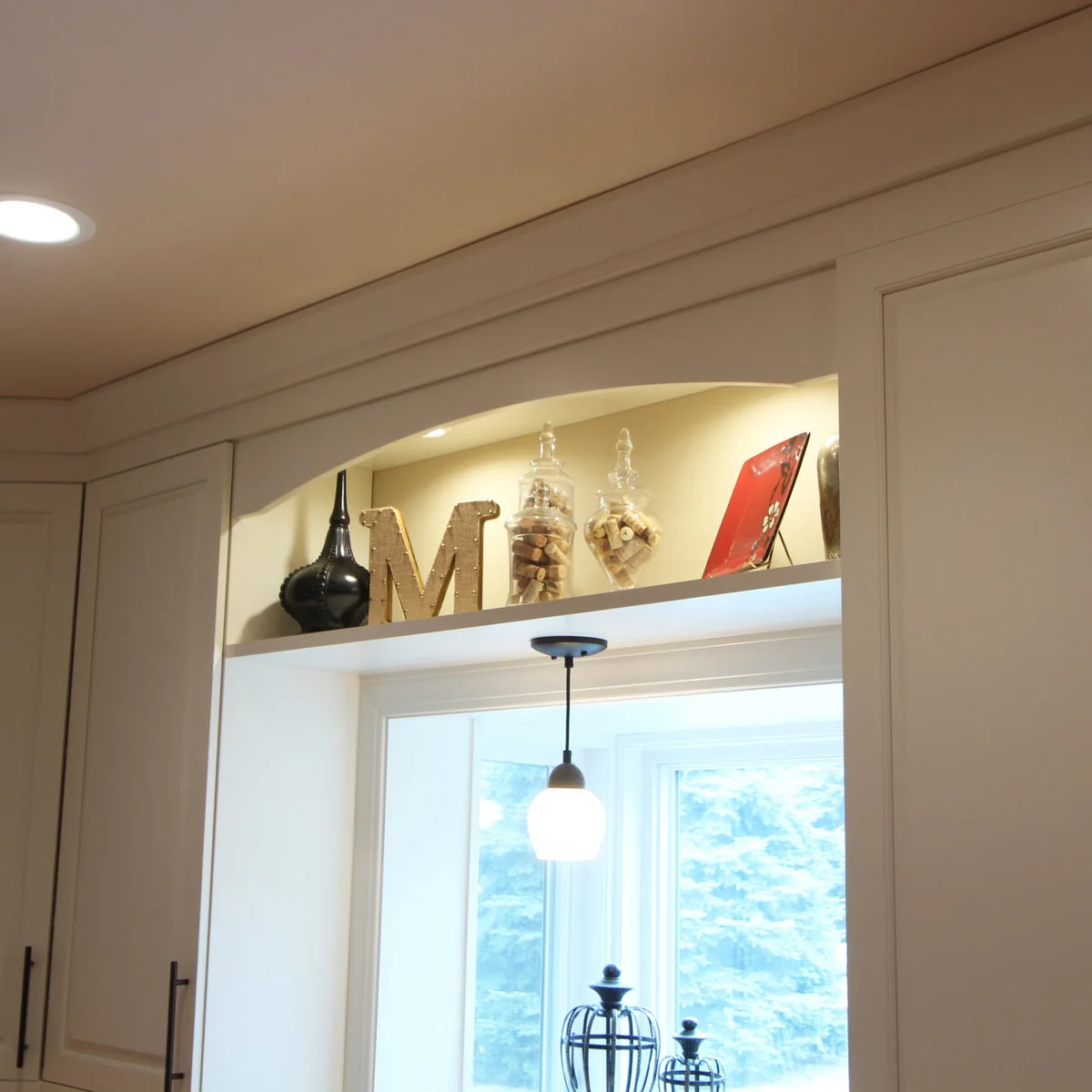A high illuminated niche above a kitchen window holds glass jars and small decorative objects, with a pendant below.