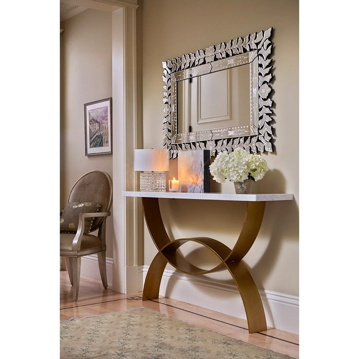 An ornate mirrored frame above a pale-topped console with a curved warm-metal base, a small lamp and white flowers.