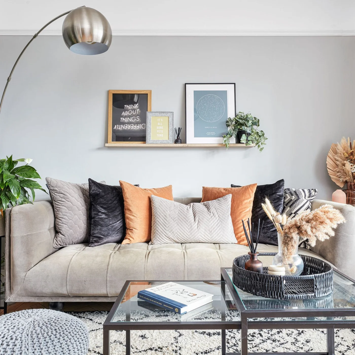 A pale sofa with rust, gray and dark cushions sits behind paired glass-topped tables beneath a narrow picture shelf.