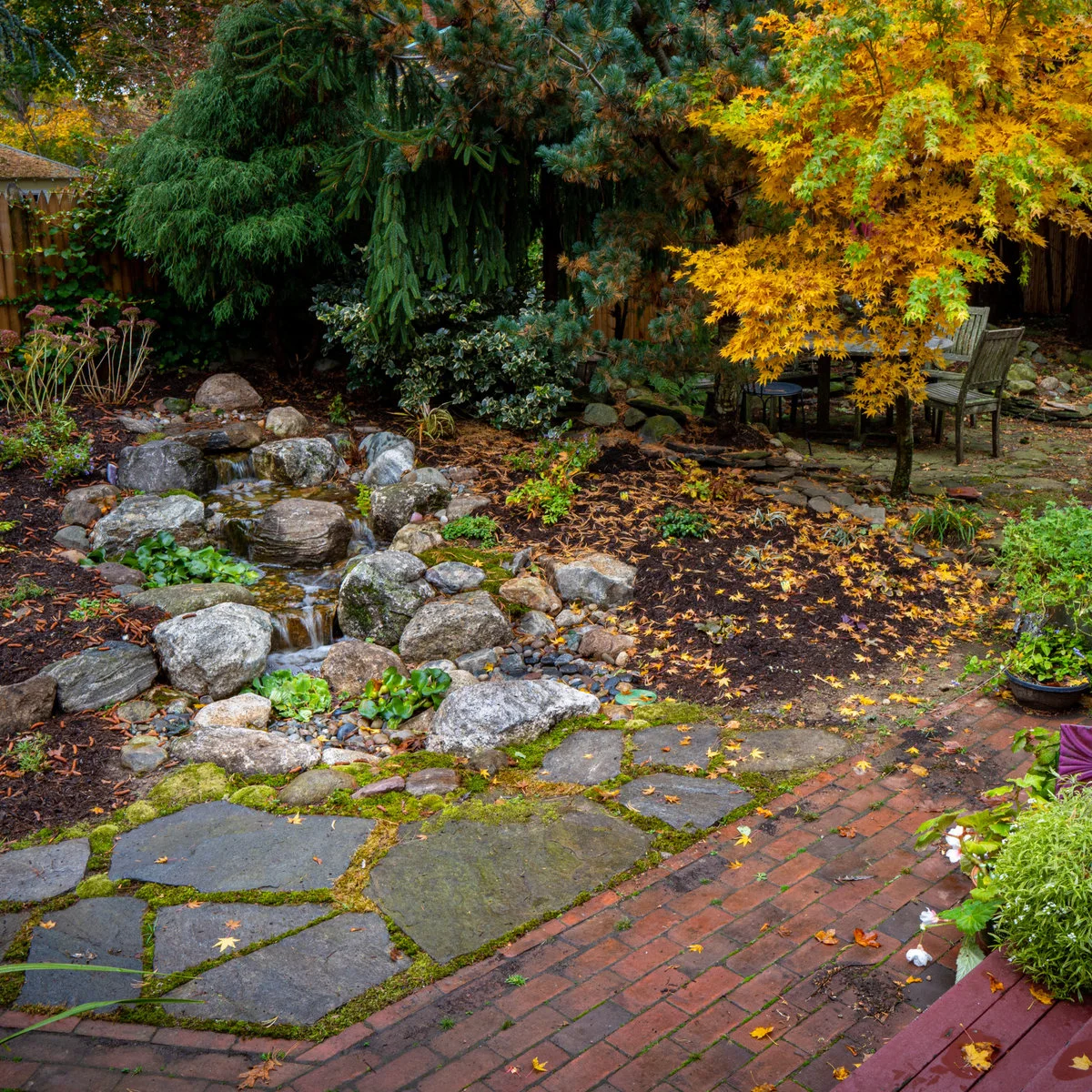 A compact rock brook beside a shaded garden dining area, with a mixed paved path, a wood fence and a yellow-leaved tree.