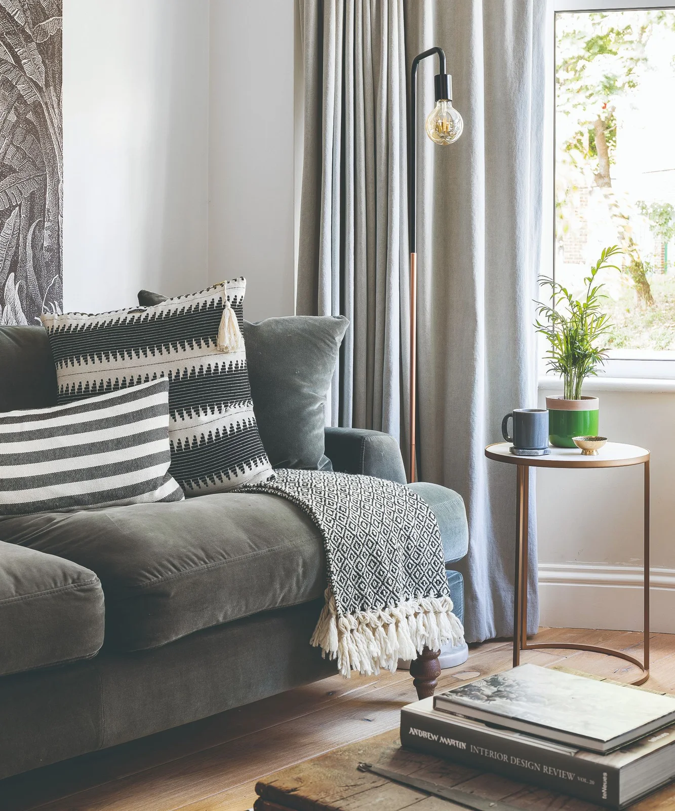 A dark gray sofa with striped cushions and a patterned throw sits beside gray curtains, a small round side table and a window