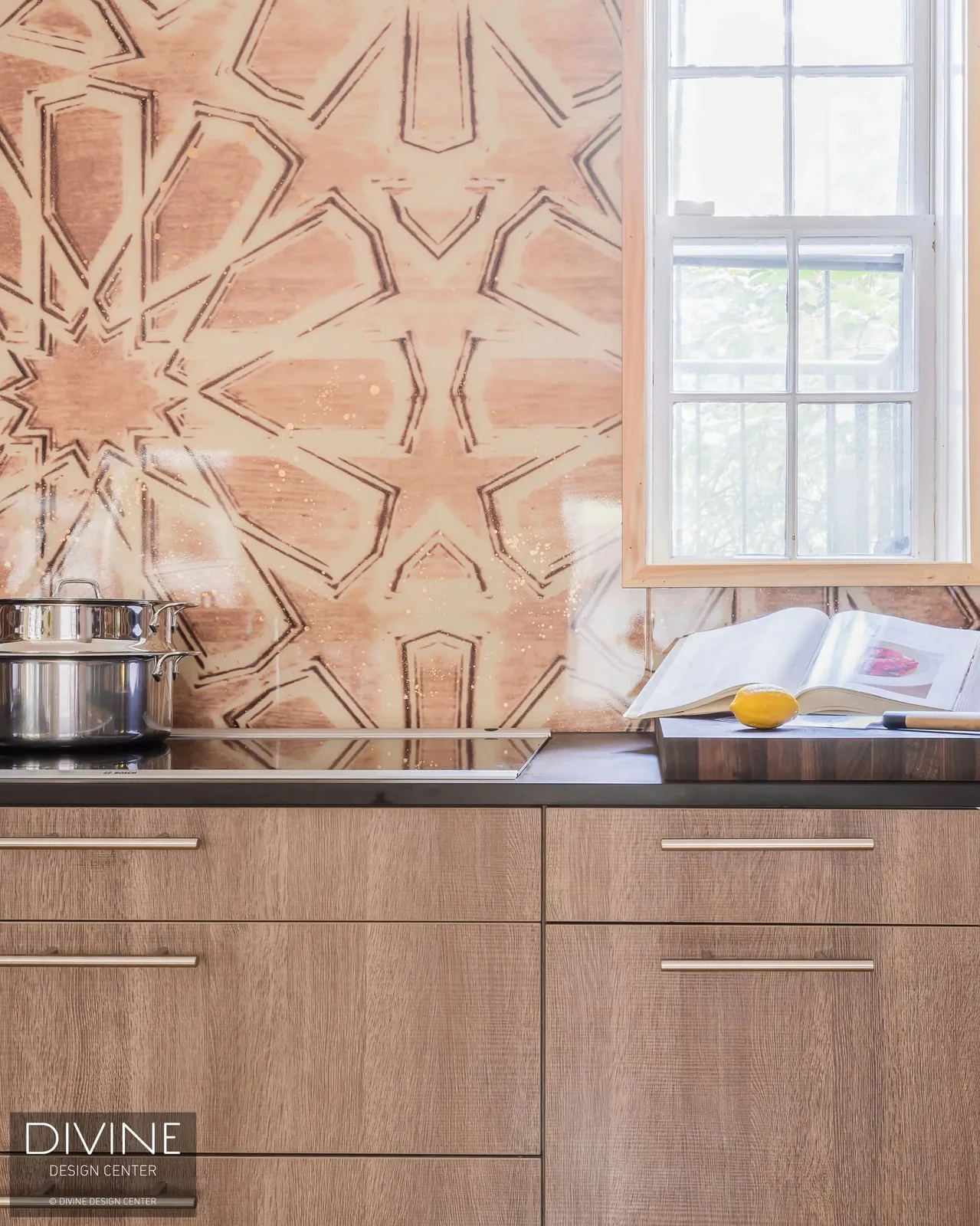 A warm star-patterned wall surrounds a white-framed kitchen window above a dark counter with a cooktop, pot, open book and wood-toned drawers