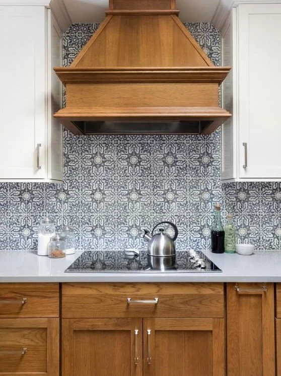 A warm wood-toned hood and lower cabinets sit between pale upper cupboards above a blue patterned cooking wall and pale counter