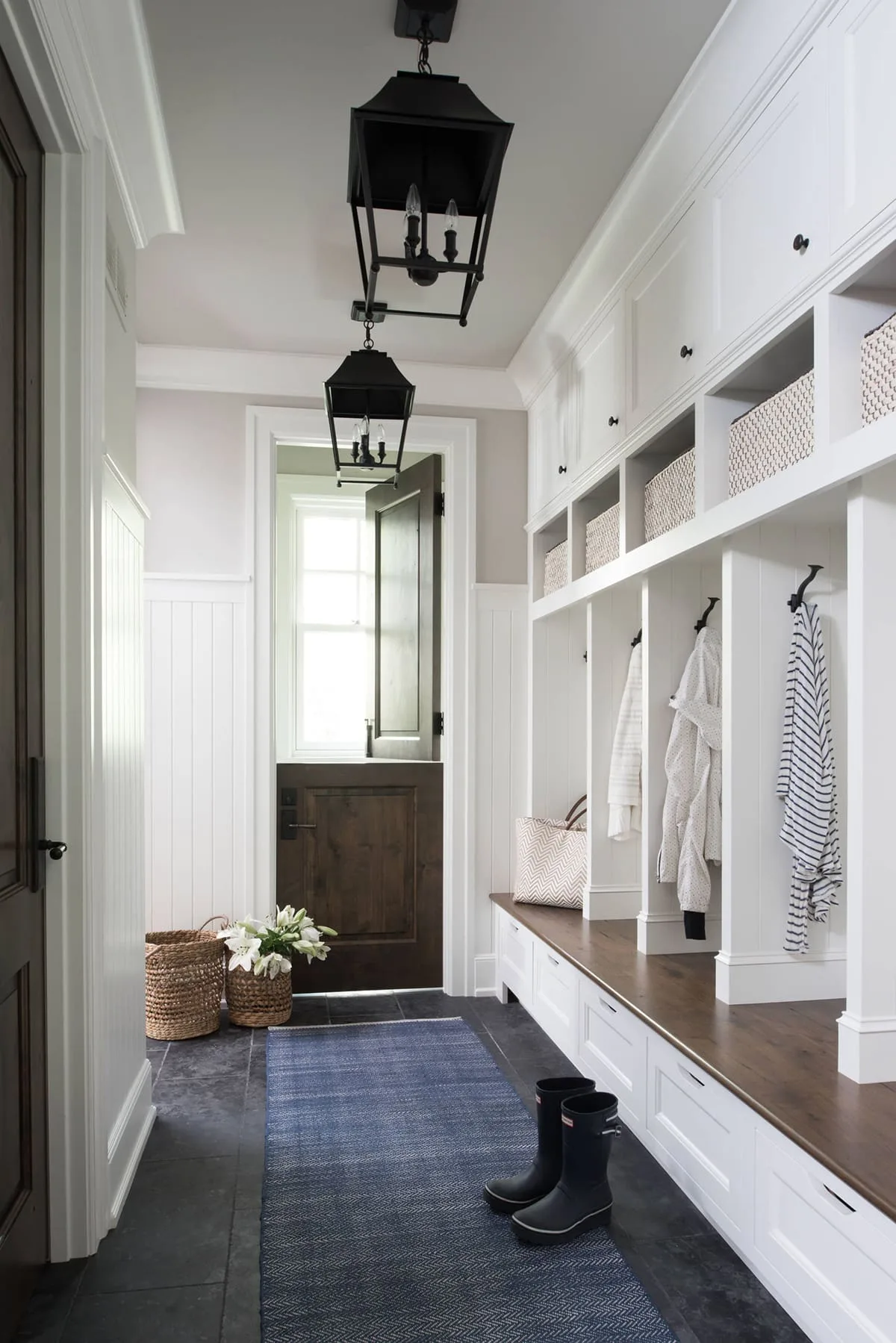 Long white mudroom storage bench with baskets, a dark divided door, two black lanterns and a blue runner.