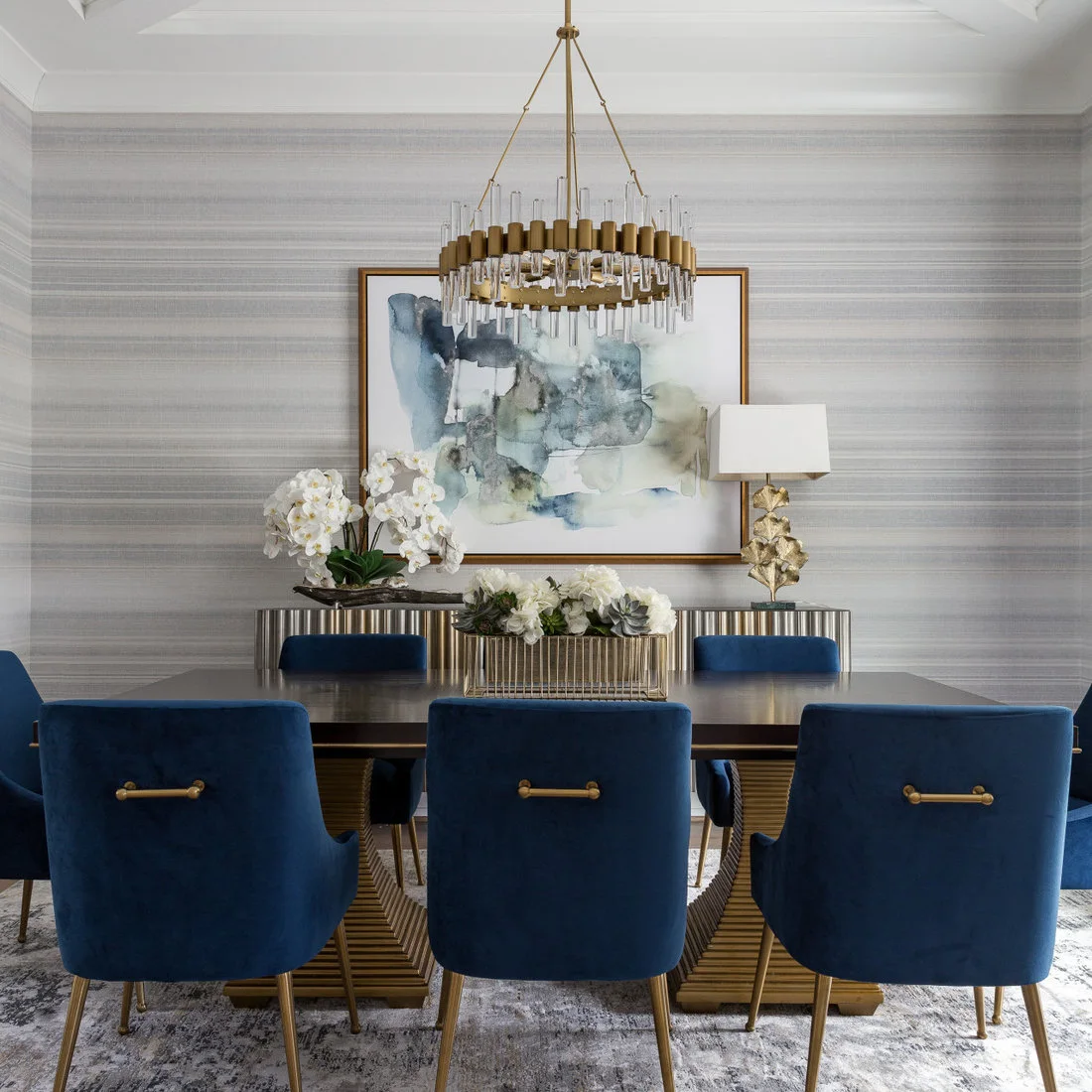 Blue upholstered dining chairs with warm-toned legs face a dark table with a geometric base beneath a circular light beside blue artwork and a ribbed sideboard