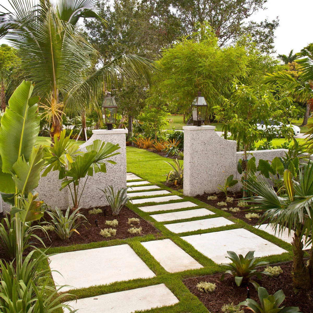 White gate pillars with lanterns frame a path of pale pavers and grass joints beside palms and broad-leaved plants.