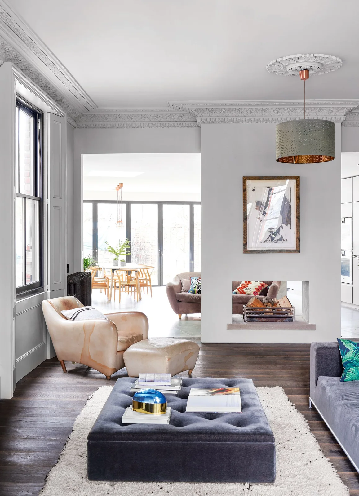 A pale room partition has a rectangular fireplace opening below framed artwork and a shaded pendant beside armchairs, a dark ottoman and an adjoining dining area