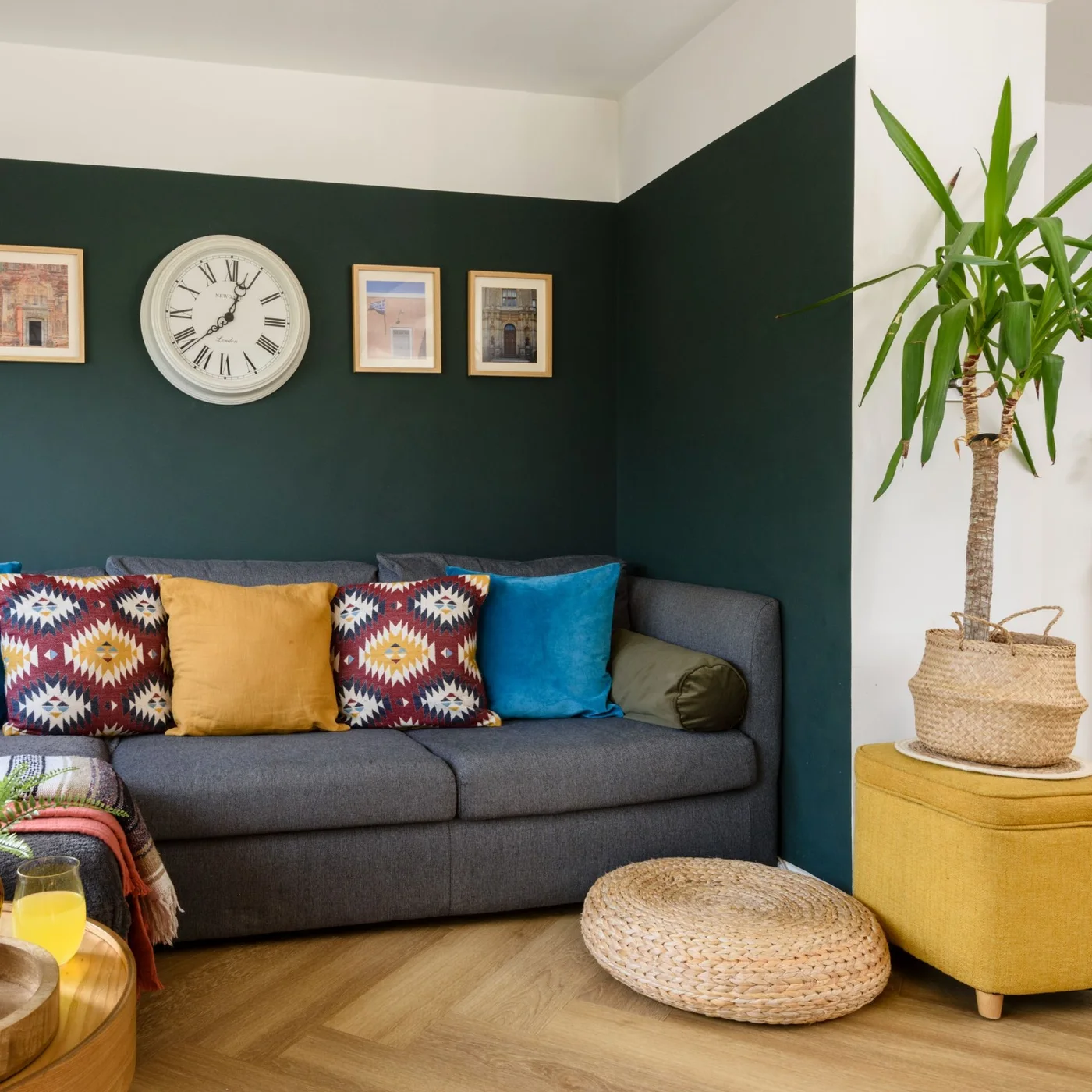 A dark gray sofa with yellow, turquoise and patterned cushions sits against deep green walls beside a mustard ottoman and woven floor cushion