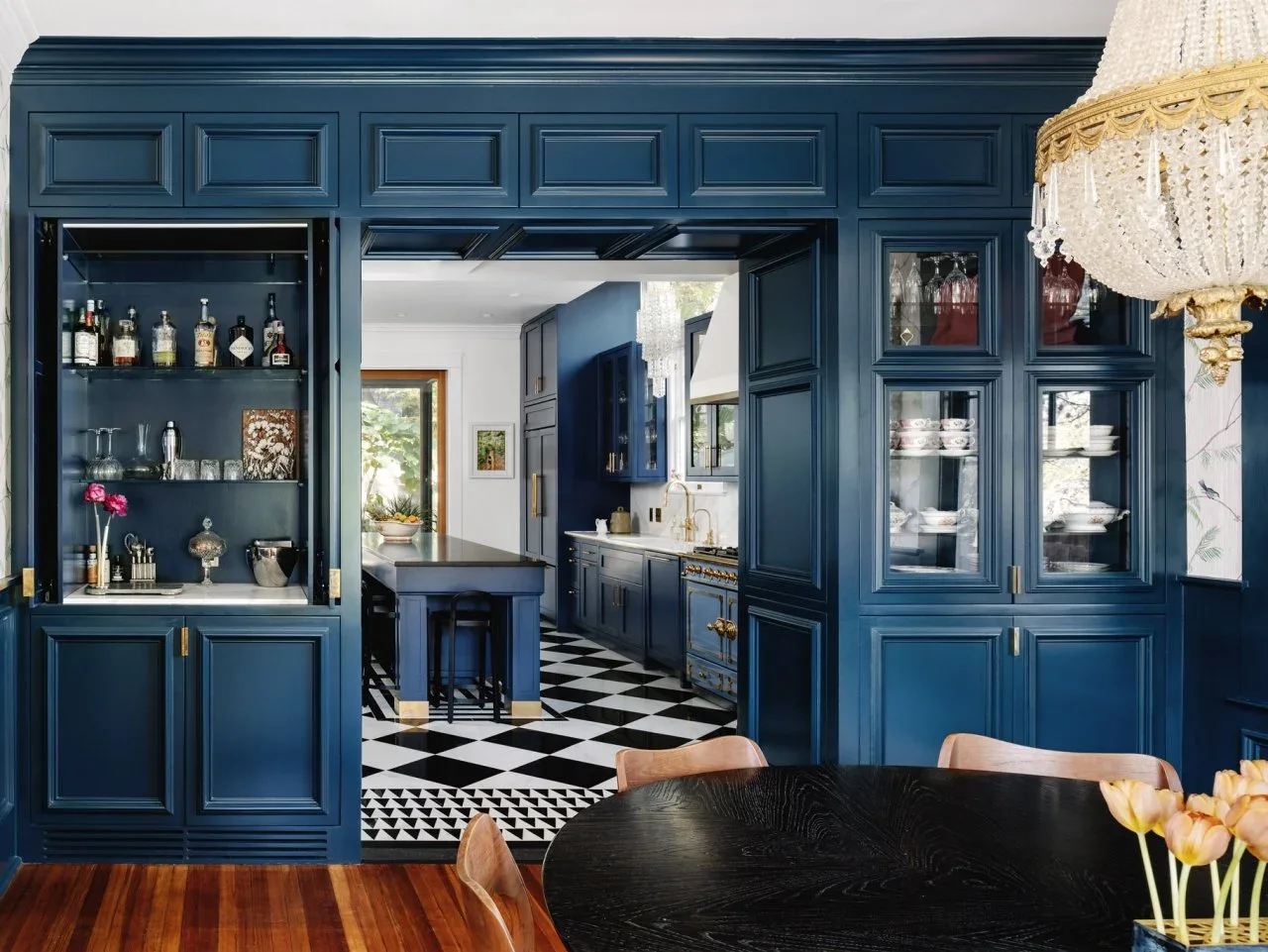 Deep blue built-in dining cabinets frame a doorway into a matching kitchen beside a dark table and timber chairs.