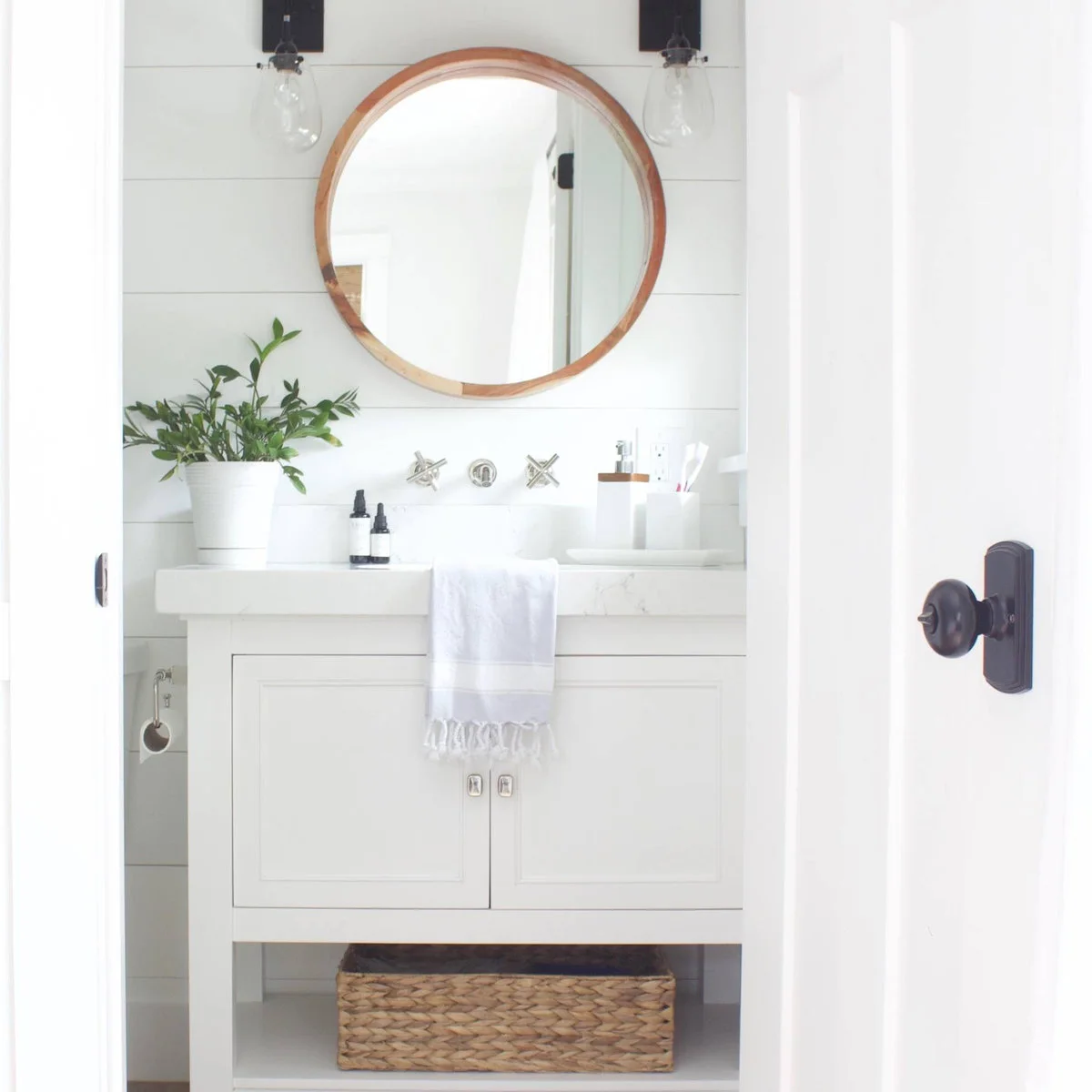 Small white bathroom vanity with a round wood-framed mirror and a woven basket on its lower shelf
