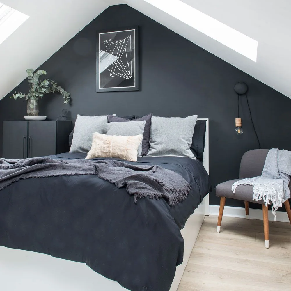Charcoal gable wall behind a gray-covered bed with white sloping ceilings and a small gray chair.