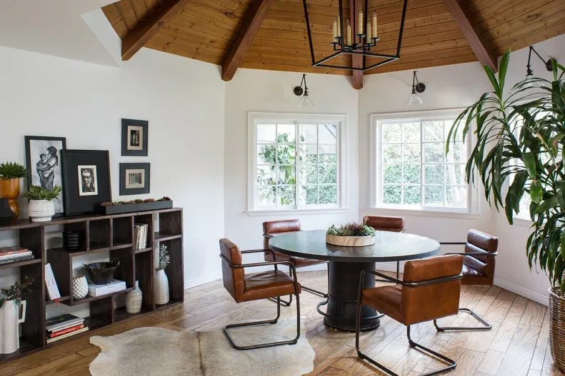 Low dark open dining bookcase with books and vessels beside a round table and brown chairs