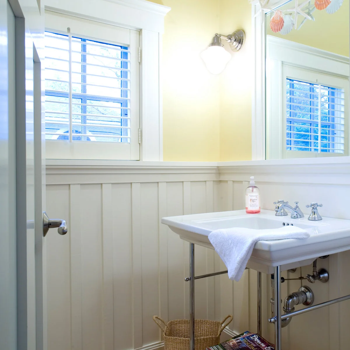 A white console sink with metal legs beneath pale yellow walls, a shuttered window and white paneling.