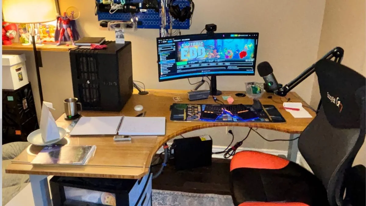 A wood gaming desk with a curved front edge, one monitor, a microphone arm, a black tower and a black-and-red chair.