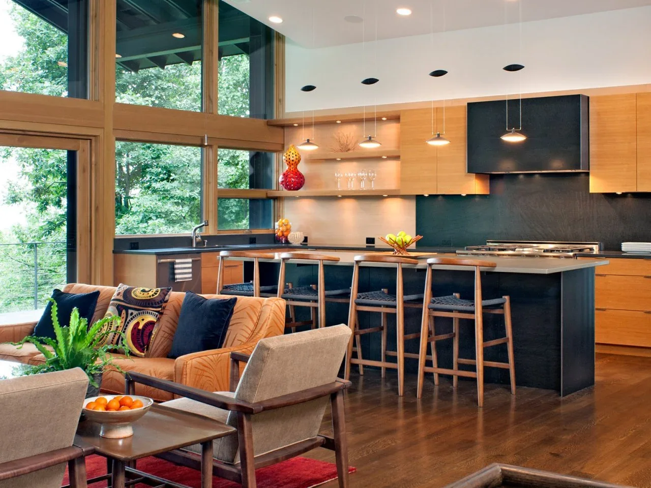 A dark kitchen island and tall stools stand behind a warm-toned sofa and timber-framed chairs beside large windows.