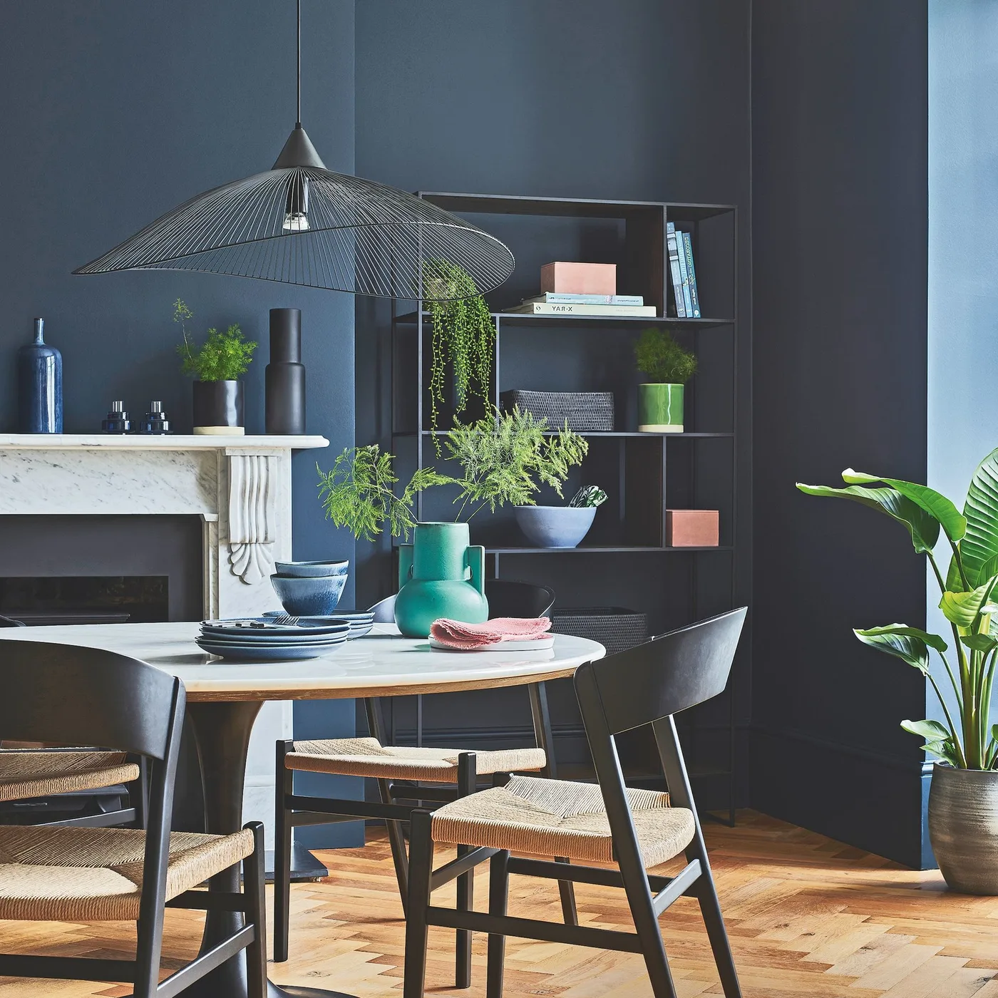 Broad open wire pendant above a round pale dining table with black chairs against navy walls