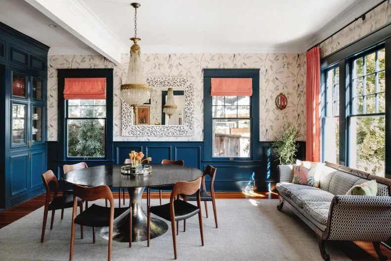 Blue dining wall panels and glazed cabinets beneath bird-patterned wallpaper, with coral blinds and curtains