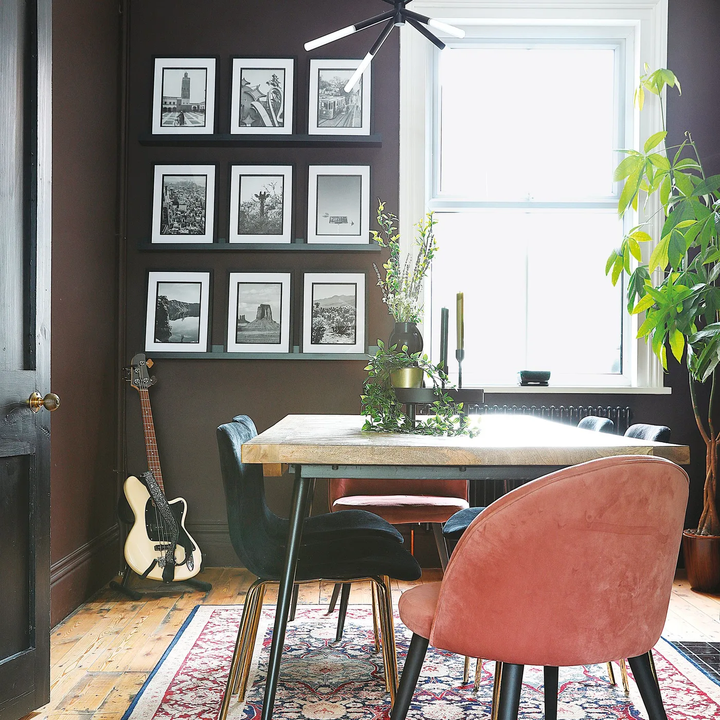 A small dining table and mixed upholstered chairs sit in a brown room with black-and-white pictures on ledges beside a large pale-framed window.