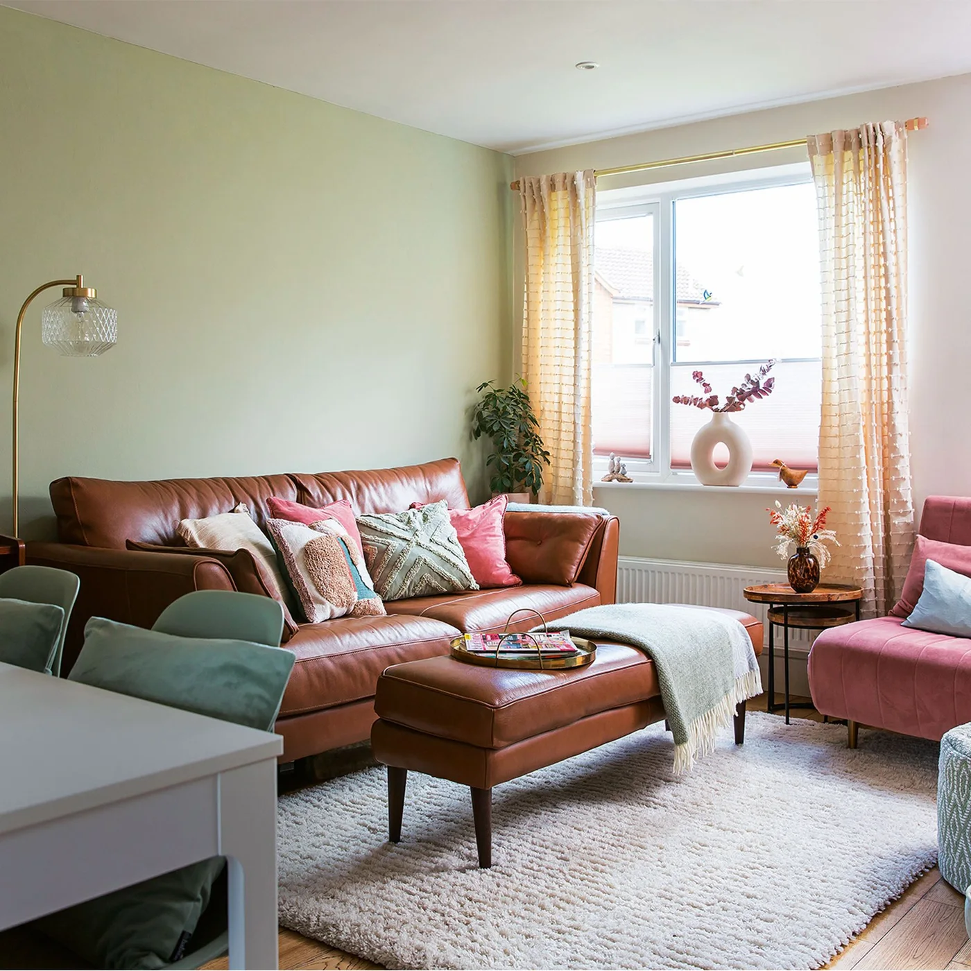 A brown leather sofa and matching footstool sit against pale green walls beside pink seating and peach curtains