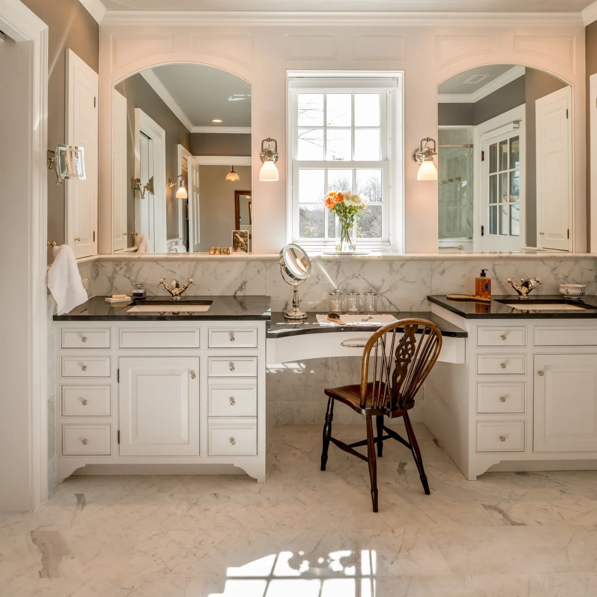 A wood chair stands at a lower dressing counter between white bathroom vanities with dark tops and tall arched mirrors.
