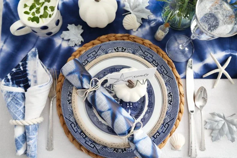 Blue-and-white plates hold a pale pumpkin and name card beside a blue napkin, woven charger and coastal decorations