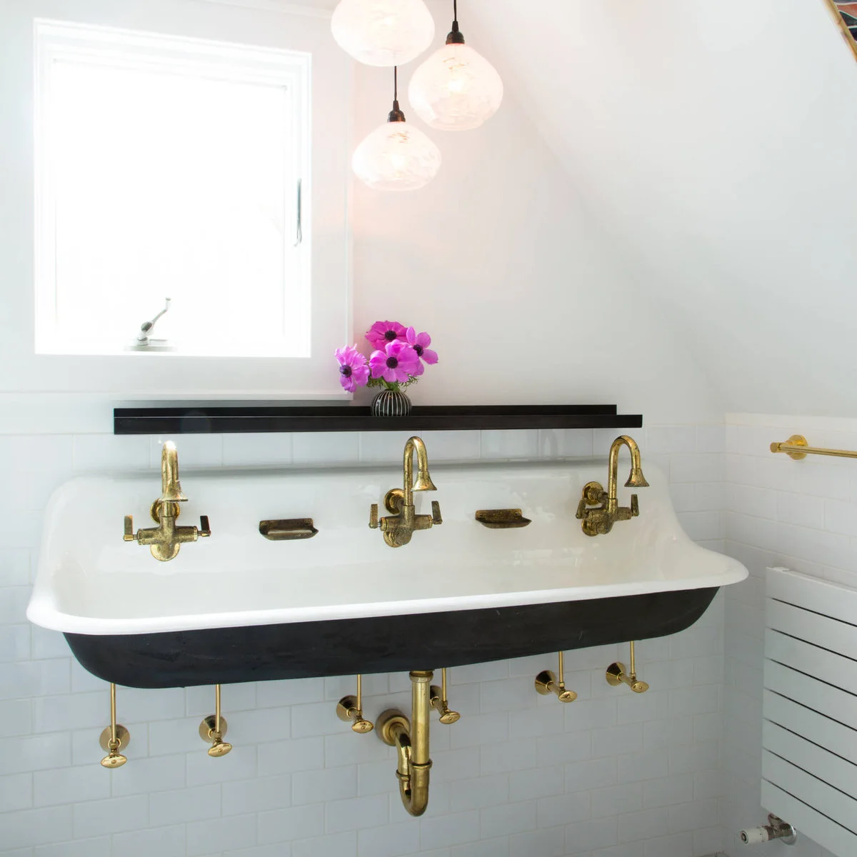 A white trough basin with a dark outside has three aged-looking gold-colored taps and exposed pipework beneath a narrow dark shelf.