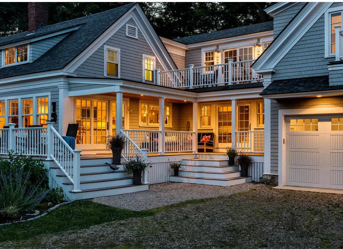 A gray shingle-sided Cape-style home has an L-shaped covered porch, white railings, two stair approaches and a balcony above.