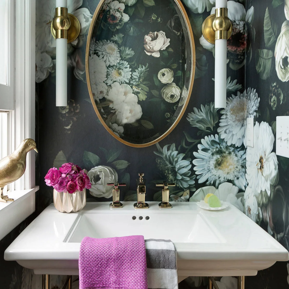 Dark floral wallpaper behind a white console basin, with an oval mirror and white cylindrical sconces.