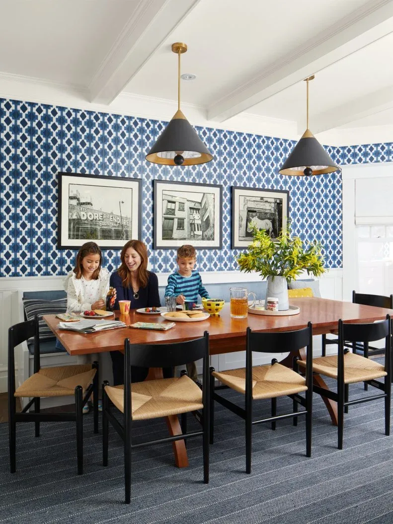 Navy-and-white geometric wallpaper sits behind a dining bench and three framed photographs, with a timber table and dark chairs.