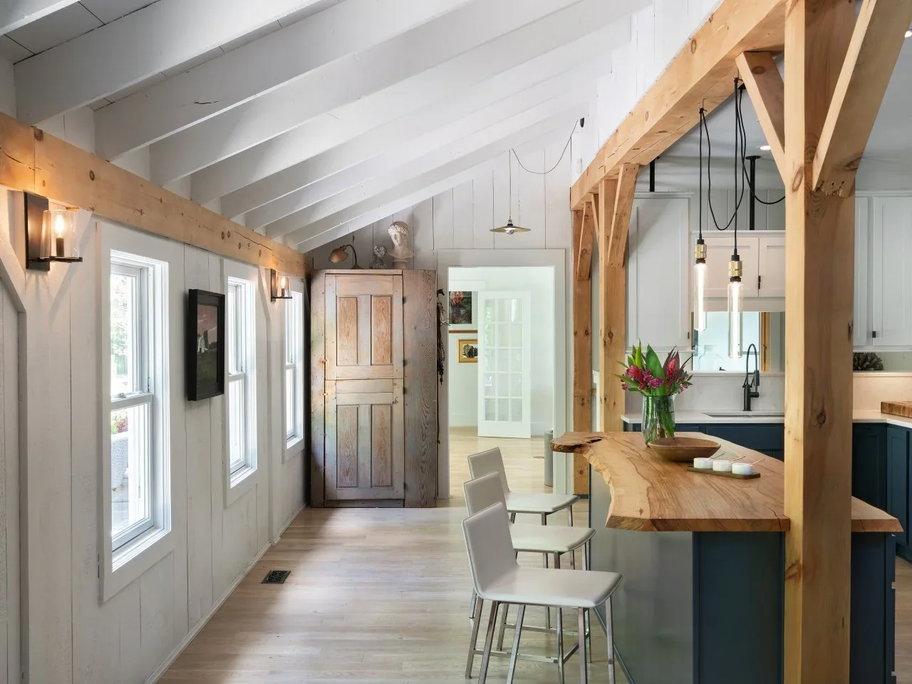 A live-edge timber breakfast counter and pale stools sit beneath exposed timber framing in a white kitchen.