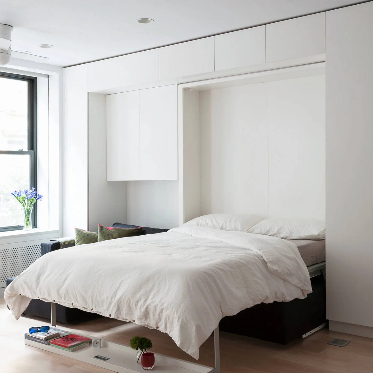 Open wall bed within full-height white cabinetry in a compact room beside a window