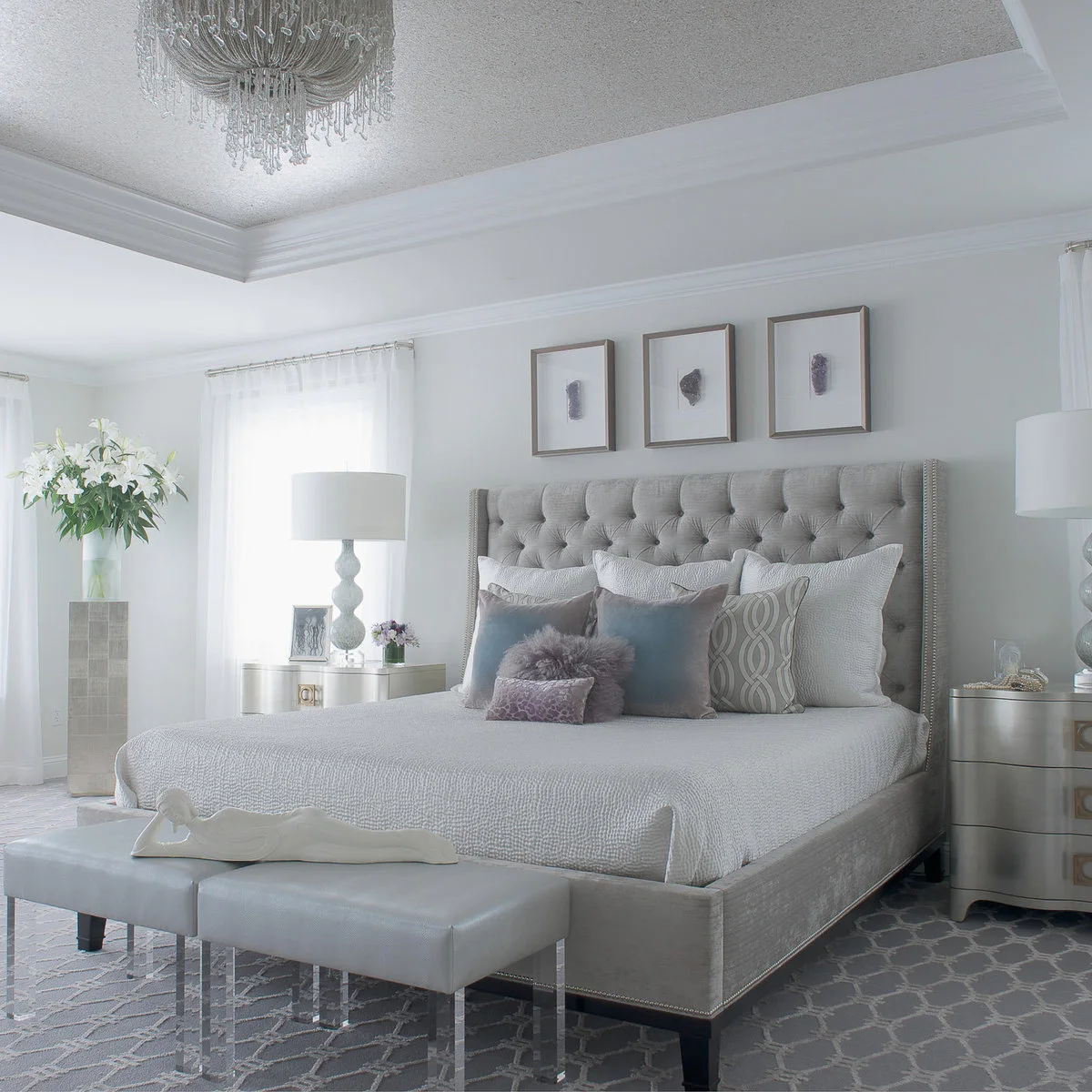 A gray tufted bed with pale bedding and several decorative pillows, flanked by curved bedside chests and two small stools.
