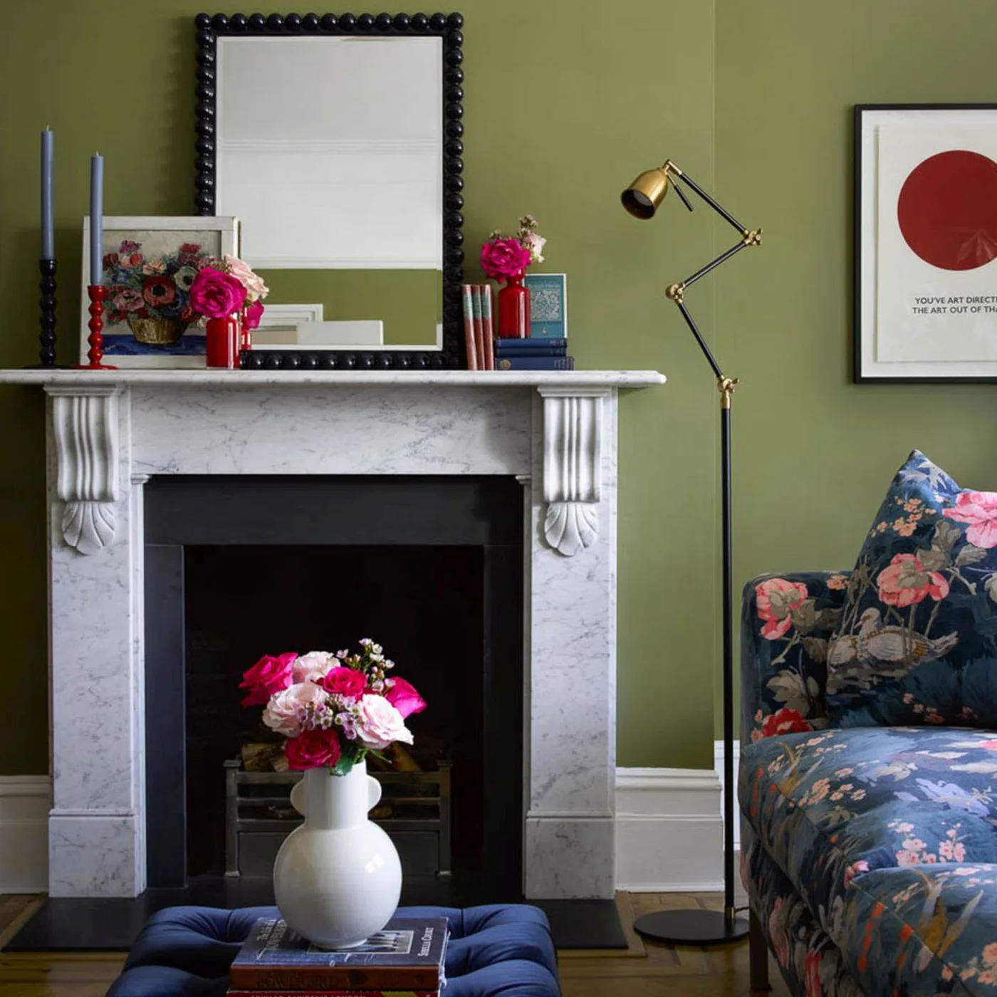 A floral sofa sits beside a pale fireplace, black beaded mirror, blue ottoman and green wall