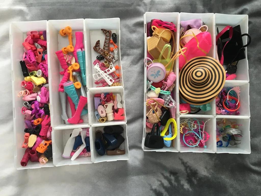 Two white divided trays holding miniature shoes, boots, handbags, hats and jewelry.
