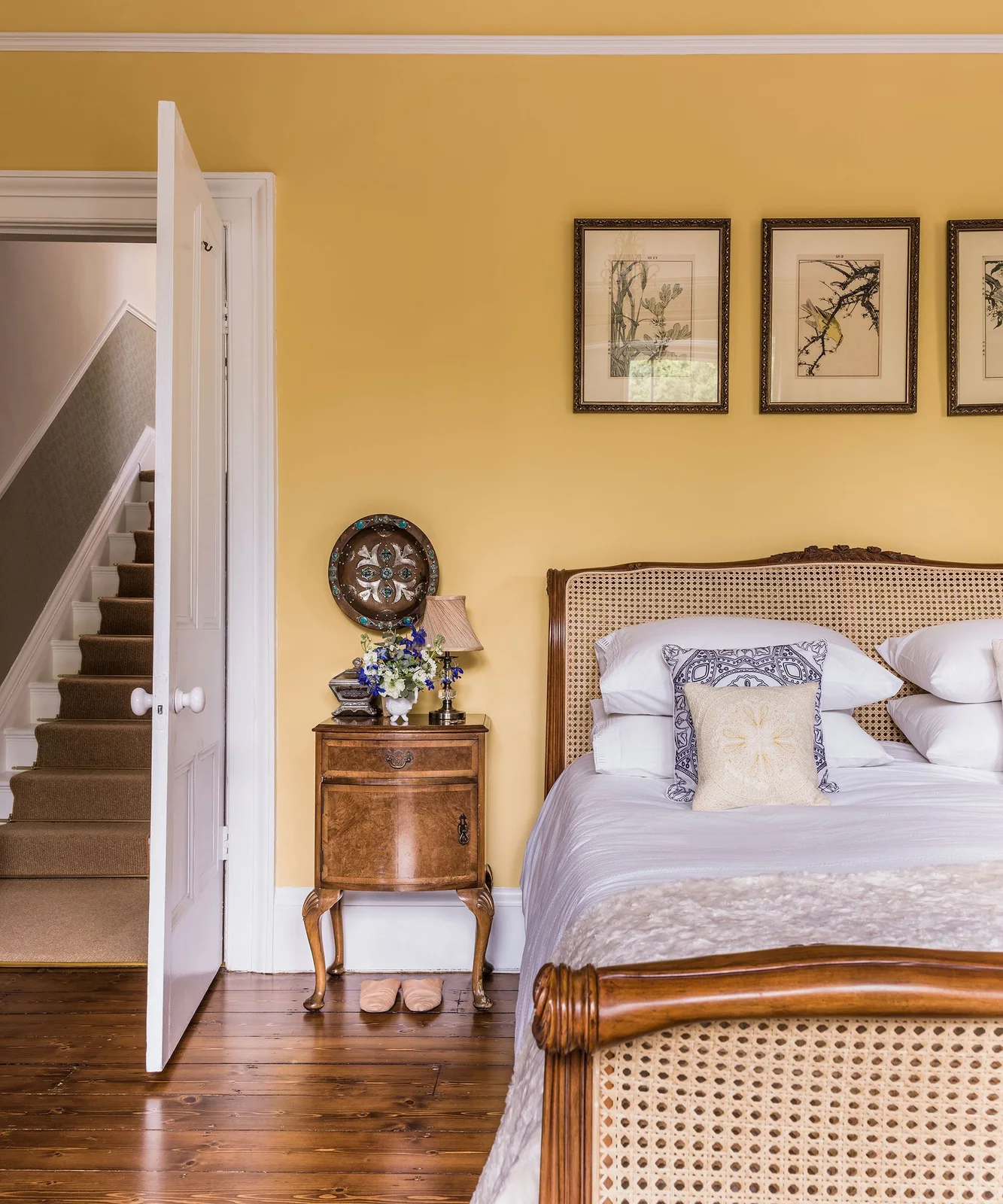 Ochre walls frame a cane-backed wood bed with white bedding, purple patterned pillows, bird pictures and a curved wood bedside cabinet.