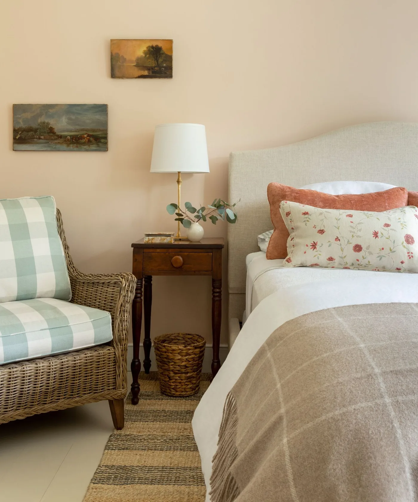 Country bedroom with a pale upholstered bed, green checked woven armchair, wooden bedside table, white lamp and small landscape paintings.