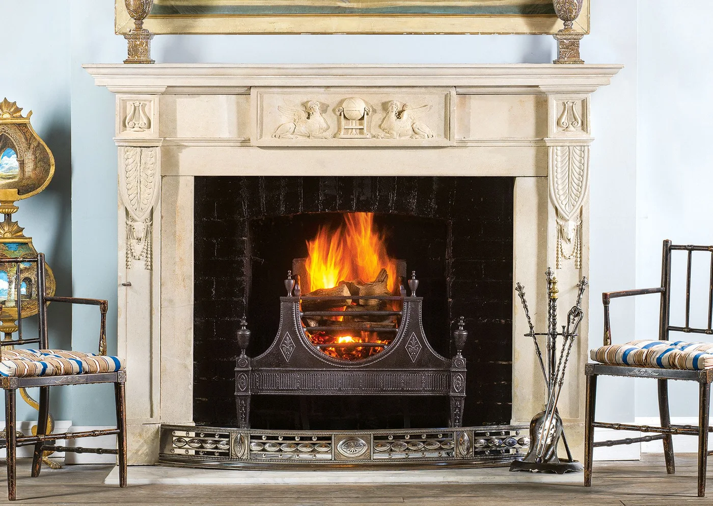 A pale carved stone mantel with relief motifs frames a dark firebox and ornate grate between slender chairs.