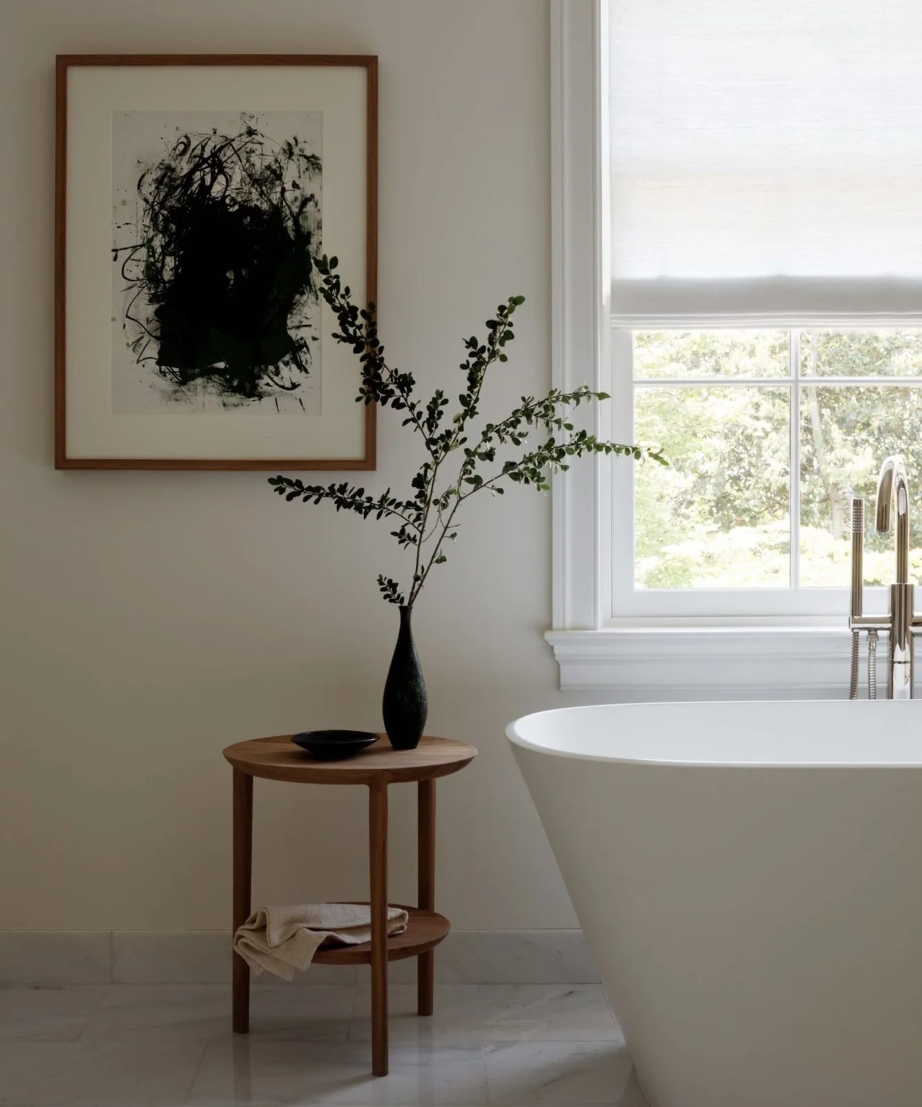 A white freestanding bathtub stands beside a two-tier round wood table with a dark vase and folded towel below framed black abstract artwork and a window