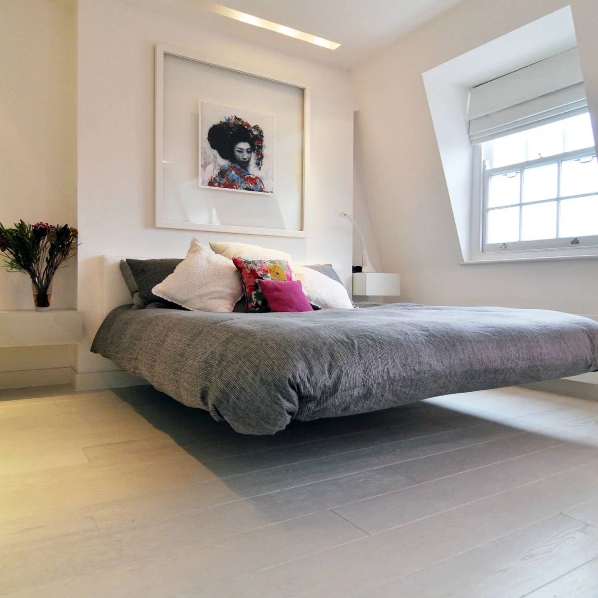 Small colorful portrait within a broad white mount above a gray-covered bed in a bright attic bedroom.