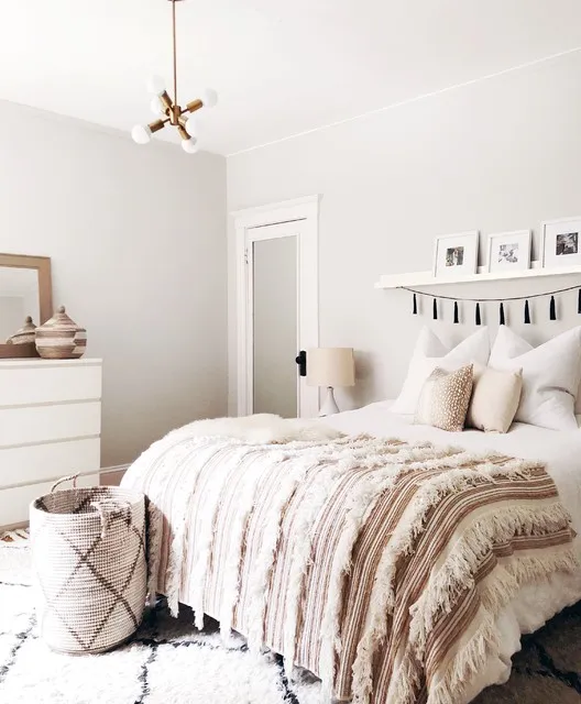 A pale bed with layered fringed bedding sits below a narrow picture ledge and small tassel trim beside a white chest and pale walls
