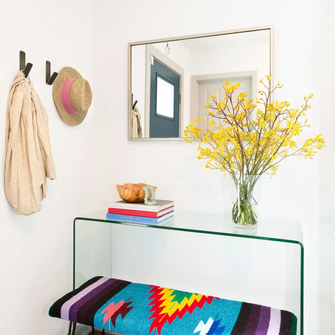 A clear glass console and rectangular mirror sit beside simple dark hooks above a bench with a bright geometric textile.